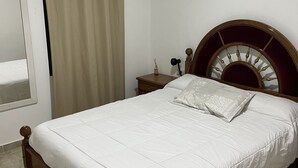 Ein ordentlich bezogenes Bett mit Holz-Kopfteil, ein kleiner Nachttisch mit Lampe und ein Fenster mit Vorhängen.