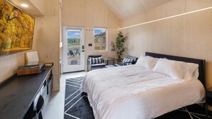 A modern bedroom with a large bed, a desk with a record player, a dining area with chairs, and a large window. - Bunkies-basecamp (Ottawa)