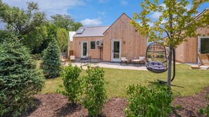 A wooden building with a sloped roof, a covered patio with outdoor furniture, and a well-maintained garden with various plants and a swing chair. - Bunkies-basecamp (Ottawa)