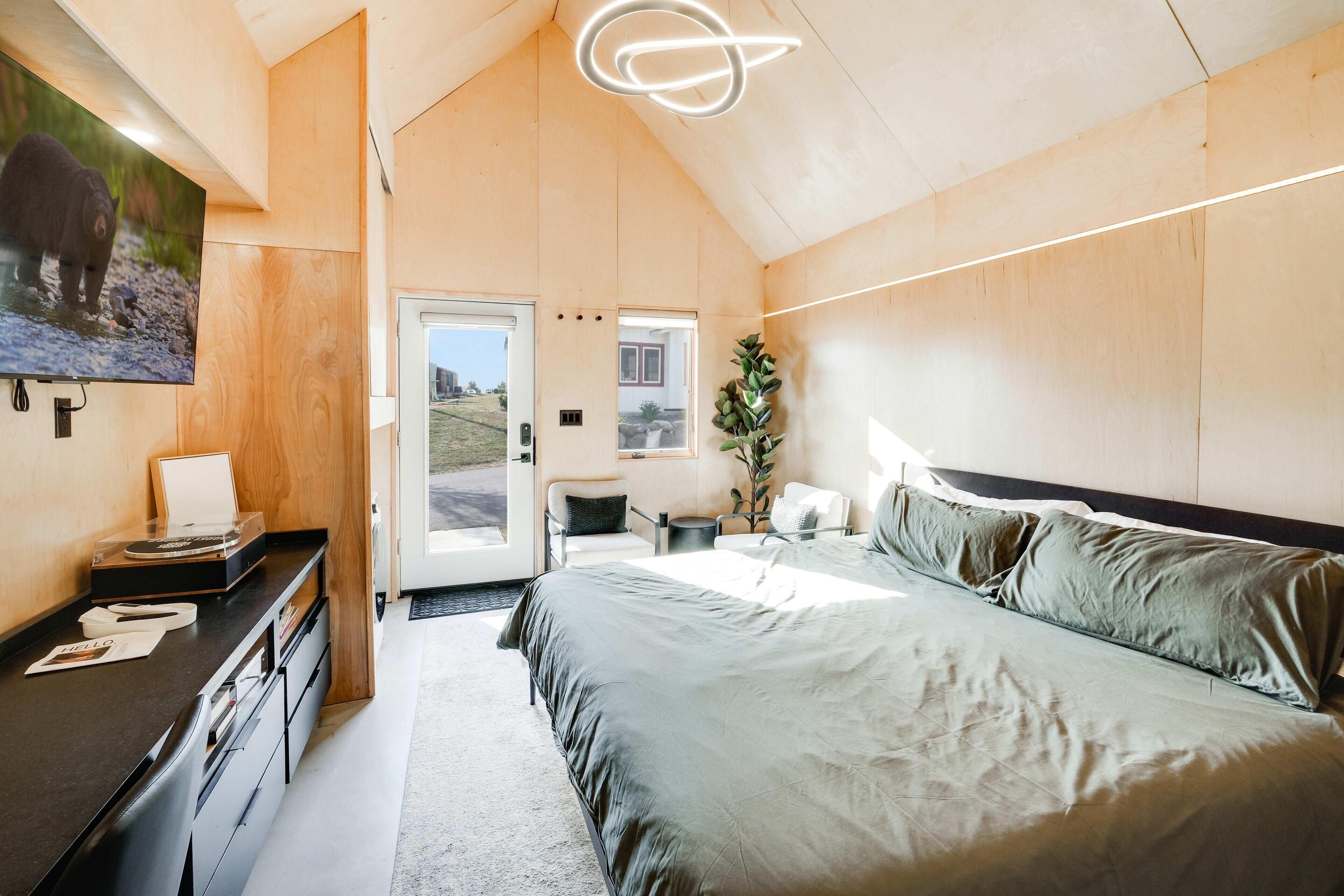 A modern bedroom with a large bed, a desk with a flat-screen TV, and a door leading to a balcony.