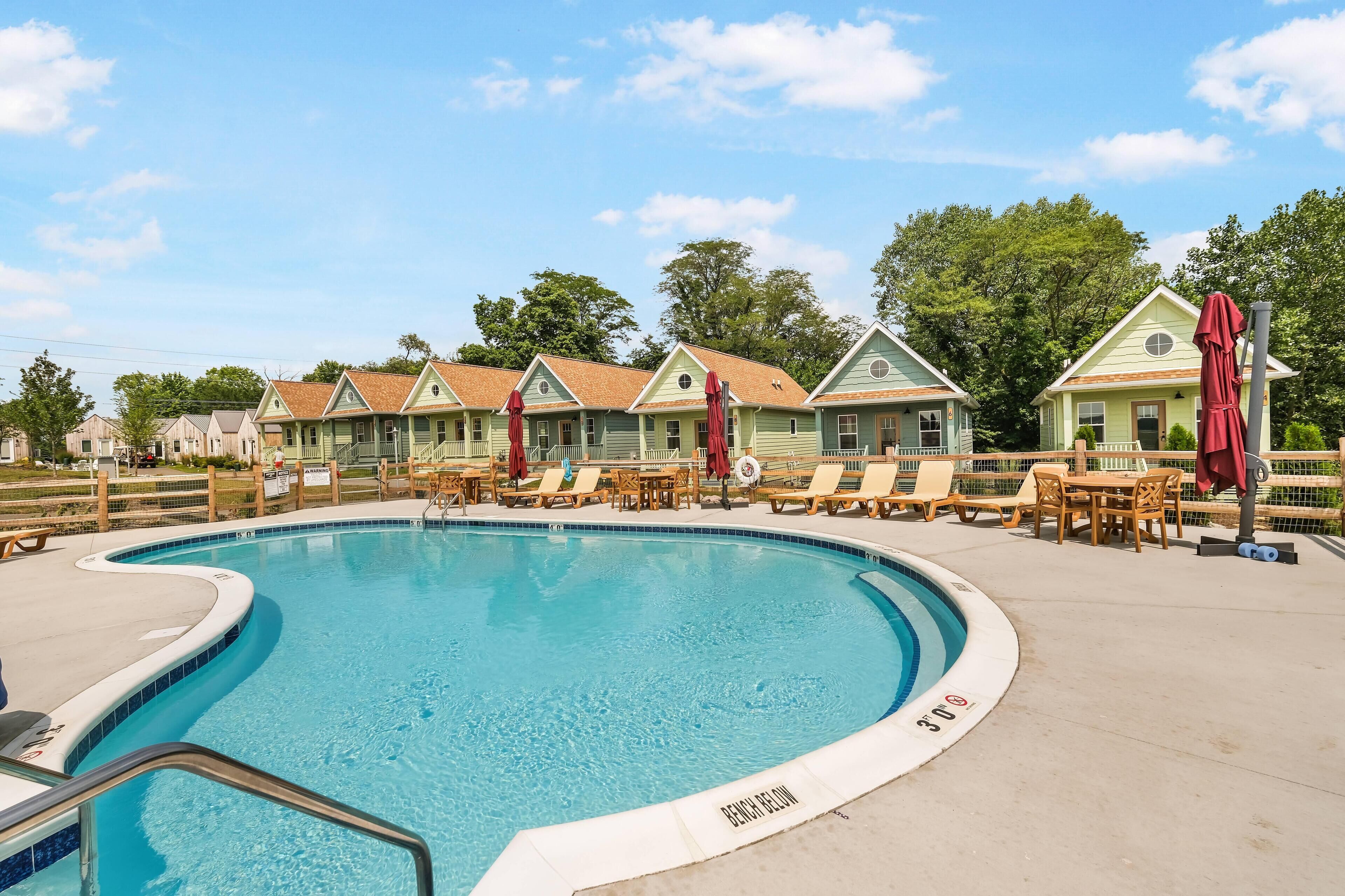 A swimming pool with lounge chairs and umbrellas, surrounded by houses with a fence.