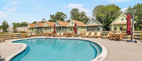 A swimming pool with lounge chairs and umbrellas, surrounded by houses with a fence.