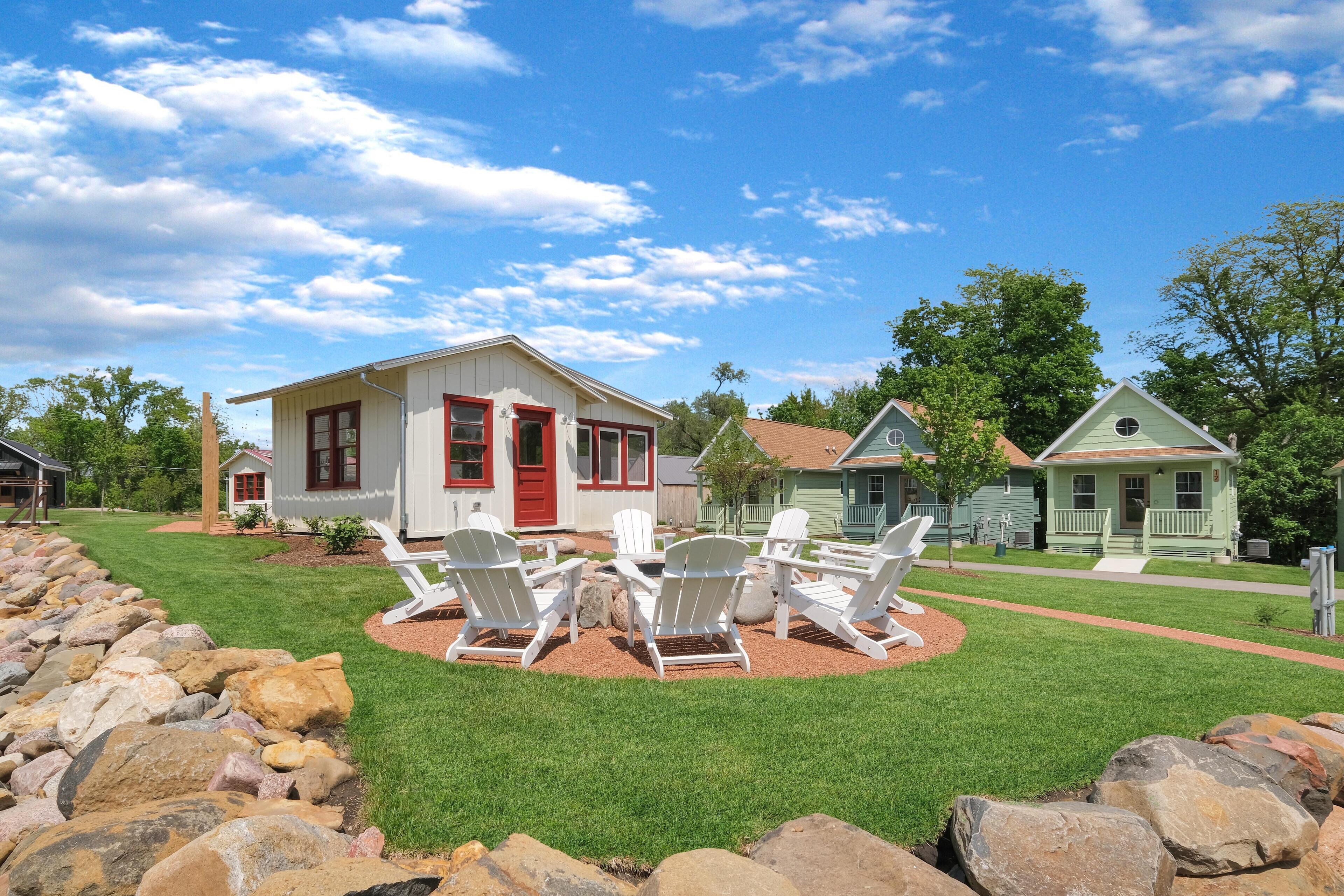 A residential area with a lawn, white Adirondack chairs, and a red door house.