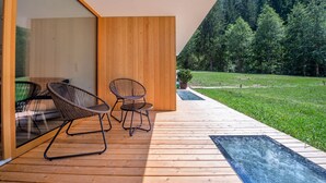 Une terrasse en bois avec deux fauteuils en osier, une porte vitrée donnant sur une pièce et une vue sur une colline boisée.