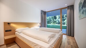 Une chambre moderne avec une tête de lit en bois, un grand lit recouvert de draps blancs et une porte-fenêtre coulissante donnant sur un balcon avec vue sur la verdure.
