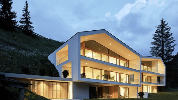 Une maison moderne à plusieurs étages, dotée de grandes fenêtres et d’un toit en pente, entourée d’une pelouse et d’arbres.