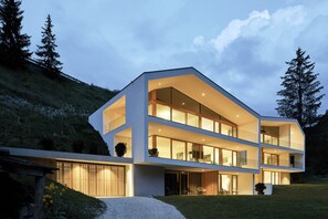 Een modern meerlaags huis met grote ramen en een schuin dak, omringd door een gazon en bomen.