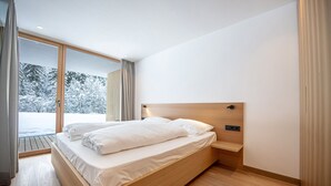 Ein modernes Schlafzimmer mit einem Holzbett, einer integrierten Nachttischschublade und einer großen Schiebeglastür, die zu einer verschneiten Außenanlage führt.