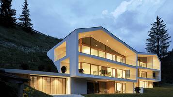 A modern multi-story house with large windows and a sloped roof, surrounded by a grassy area and trees.