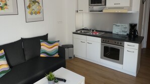 A compact kitchen with white cabinets, a microwave, a stove, and a coffee maker. There is a black sofa with colorful pillows and a small plant on a white table. - Idyllic location on the Wennsee and 10 min walk to the beach with beautiful garden. (Scharbeutz)