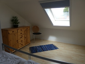 Quarto com teto inclinado, claraboia, cómoda de madeira, cadeira, tapete azul e uma cama.