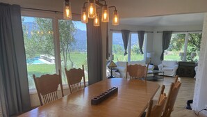 Ein Essbereich mit einem Holztisch und Stühlen, einer Kristallleuchter und großen, mit Vorhängen versehenen Fenstern.