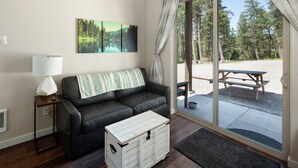 Una habitación acogedora con un sofá de cuero negro, un cofre de madera blanca, una lámpara y un cuadro de un bosque.