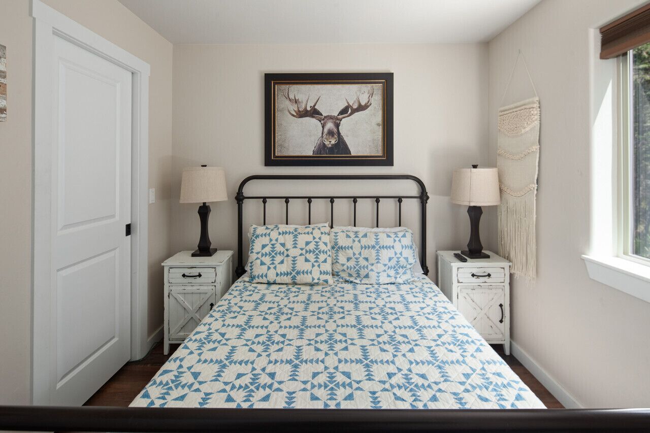 Een slaapkamer met een bed, twee nachtkastjes, een lamp op elk kastje, een hertenfoto in een lijst boven het bed en een raam met uitzicht.
