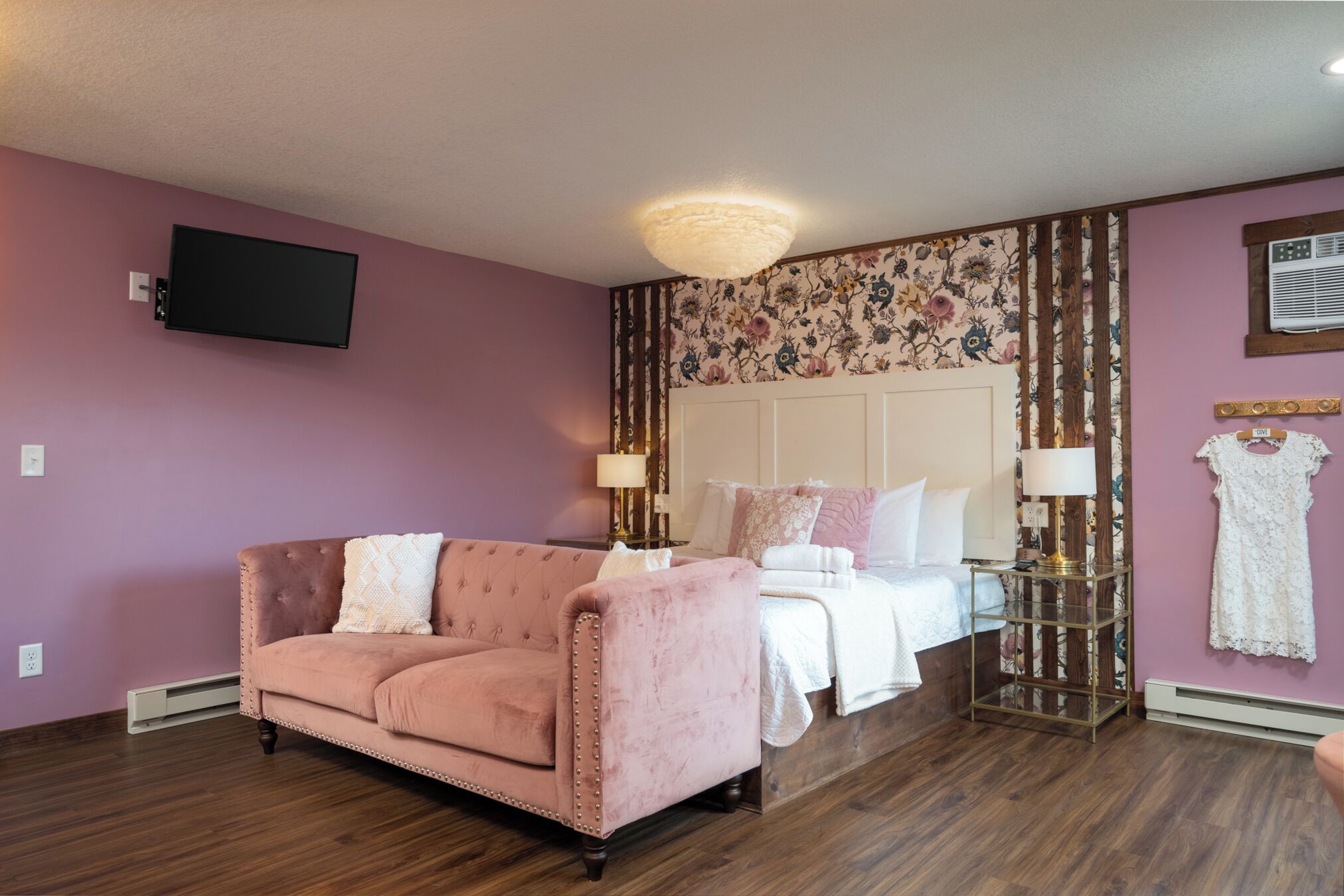 Una stanza con un divano rosa, un letto con biancheria bianca, una TV fissata al muro e una carta da parati a motivi floreali.