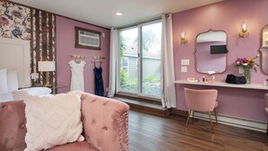 Ein Zimmer mit rosa Wand, einem großen Fenster, einem Schminktisch mit Spiegel und einem rosafarbenen Stuhl.