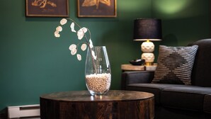 Een kamer met een ronde houten tafel, een glazen vaas met witte bloemen, een donkergroene muur en een zwarte lamp.
