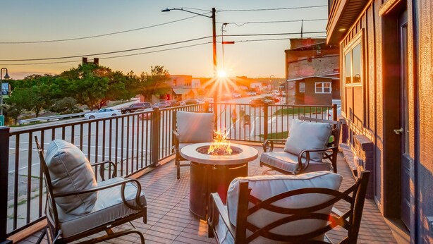 En terrass med utomhusmöbler, en eldstad och utsikt över solnedgången över ett bostadsområde.