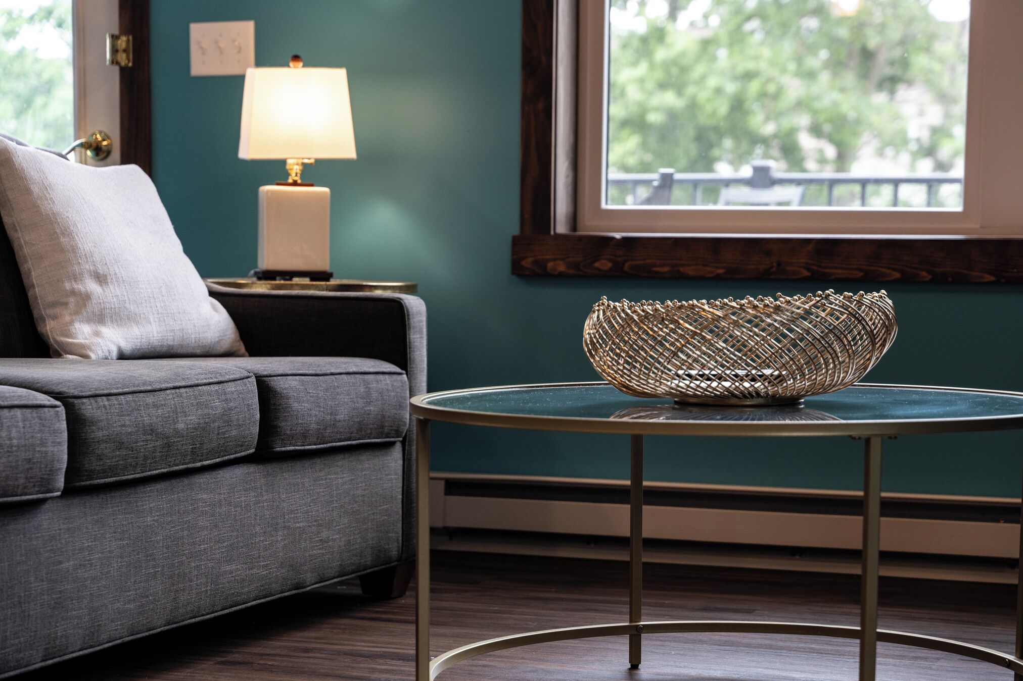 A living room with a grey sofa, a round glass table with a woven basket, a lamp, and a window with a view of greenery.
