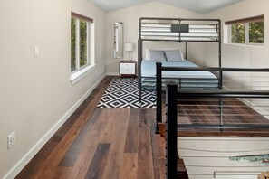 A bedroom with a bunk bed, a bedside table with a lamp, a patterned rug, and a window with a view of trees. - Swift Current at 406 Glacier Cabins (Columbia Falls)