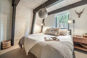 A cozy bedroom with a large bed, a hanging light fixture, a deer head wall decoration, and a window with a view of trees. - Northstar A-Frame Cabin with Hot Tub | Resort Shuttle, Sleeps 8 (Truckee)
