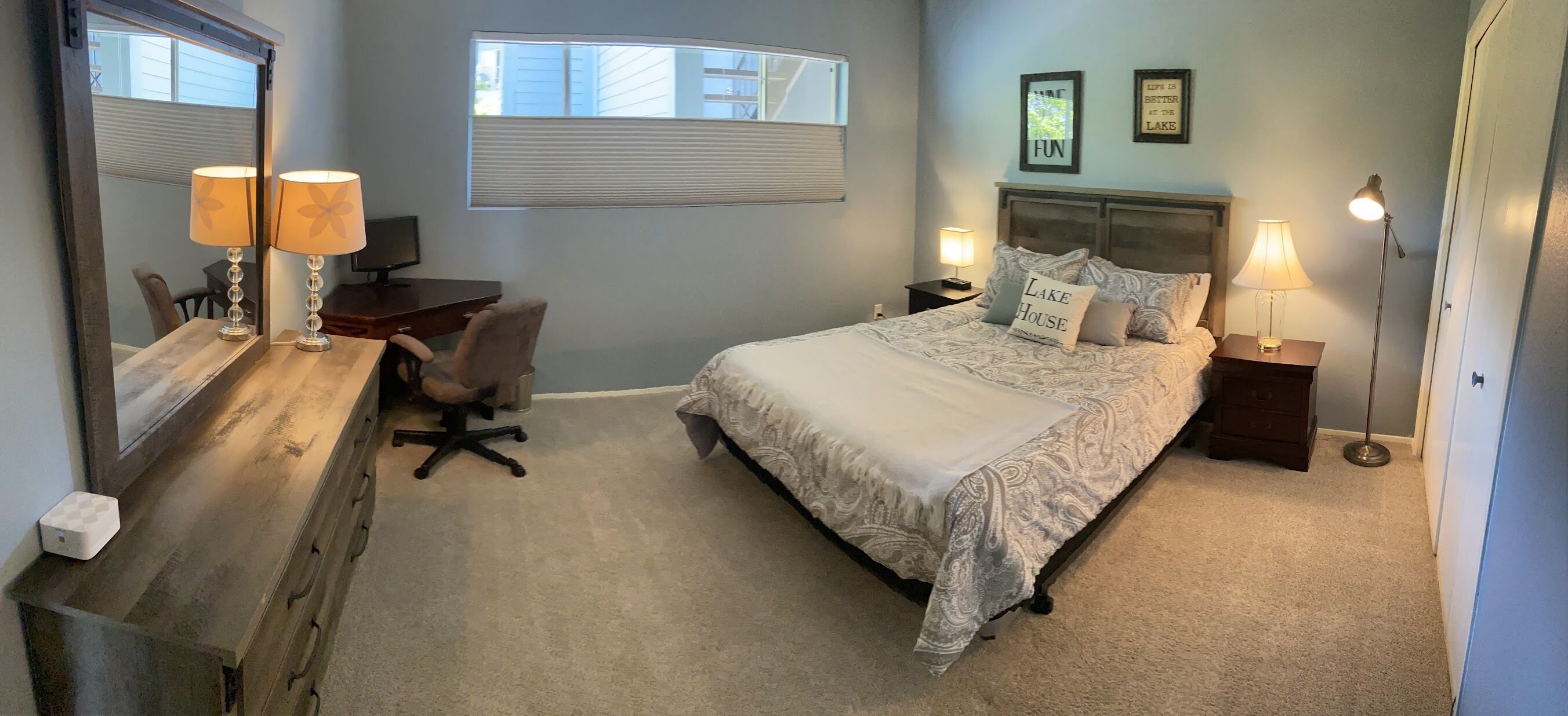 Une chambre à coucher comprenant un lit, deux tables de chevet avec des lampes, un bureau avec une chaise, un grand miroir et une fenêtre avec des stores.