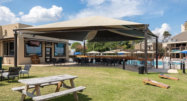 Un espace extérieur aménagé en terrasse, doté d’un grand auvent, de tables de pique-nique et d’une piscine en arrière-plan.