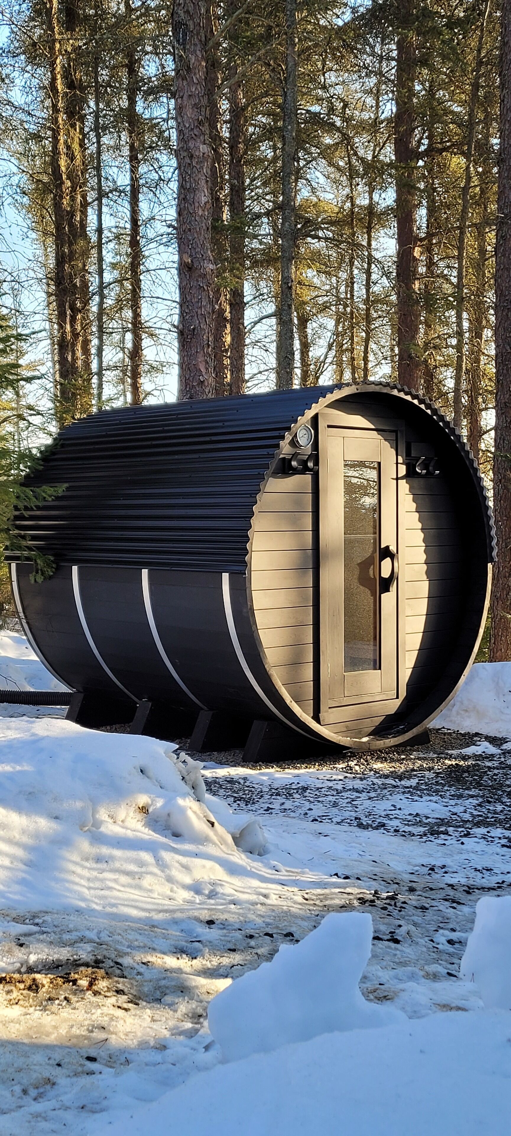 Une cabane cylindrique en bois, dotée d’une porte, entourée de neige et d’arbres.