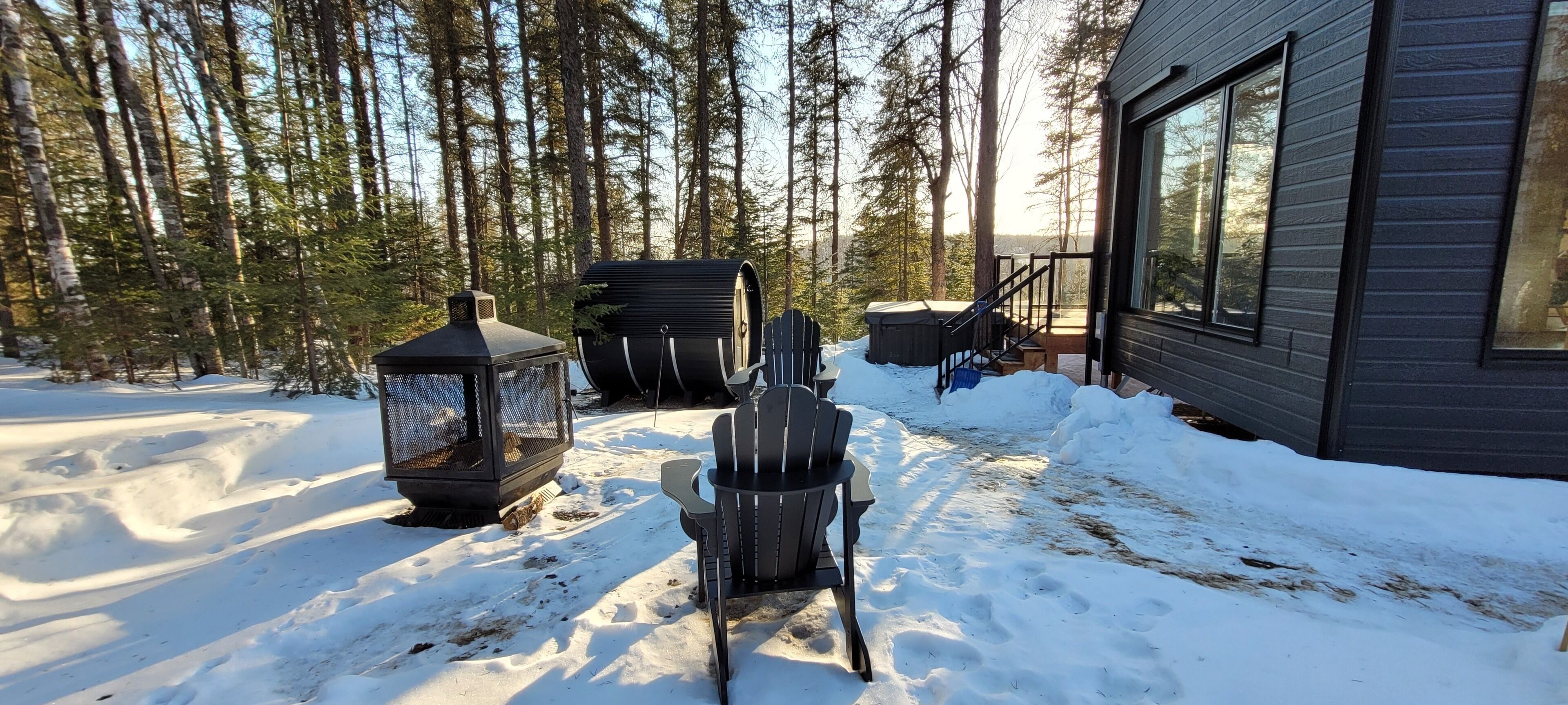 Un espacio exterior cubierto de nieve con una terraza de madera, una silla negra, un comedero para pájaros y un jacuzzi.