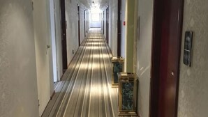 Un long couloir étroit, typique des hôtels, avec des portes de chaque côté, un sol recouvert de moquette et des colonnes surmontées de marbre.
