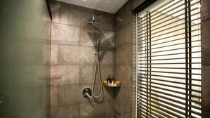 Une douche équipée d’un pommeau de douche à main et d’un robinet mural, une étagère pour les articles de toilette et une fenêtre avec des stores.