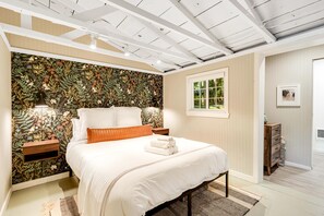 Un dormitorio con papel tapiz floral, una cama con ropa de cama blanca, una mesita de noche y una ventana.
