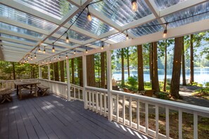Una terraza cubierta con mobiliario de madera y luces colgantes, con vistas a un lago rodeado de árboles.