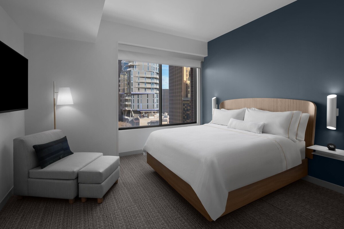 Ein modernes Hotelzimmer mit einem großen Bett, einer Sitzecke, einem Fernseher und einem Fenster mit Blick auf die Stadt.