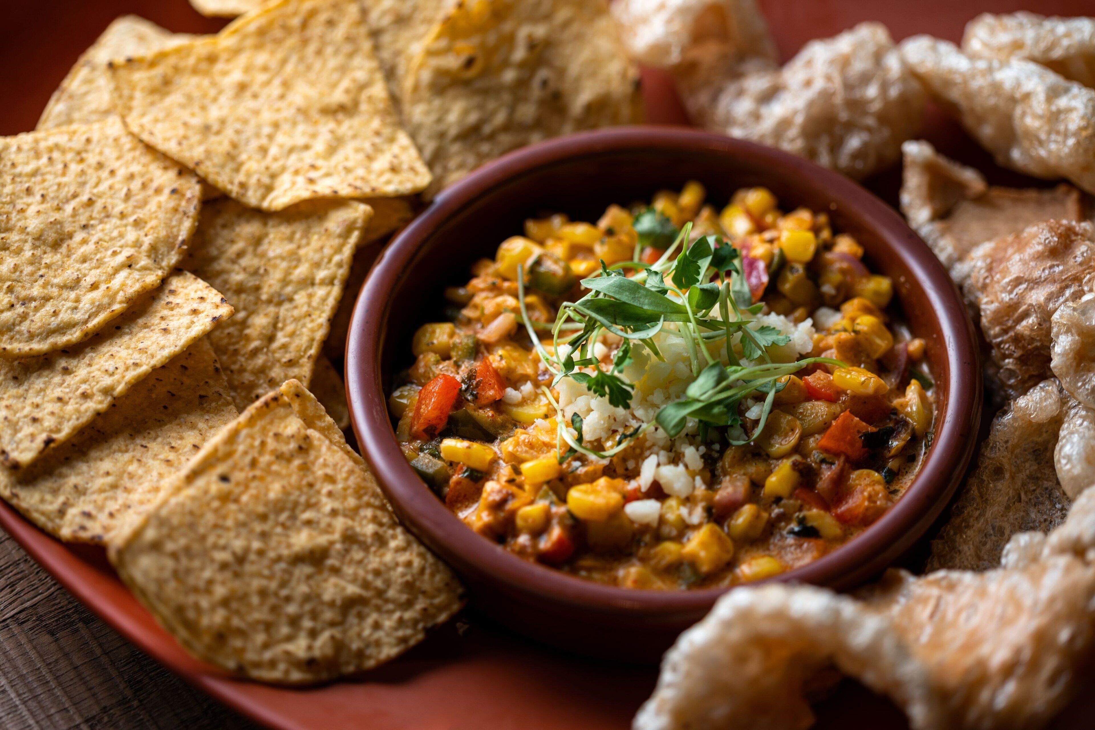 En skål grøntsagsgryde med majs, gulerødder og bønner, serveret med tortillachips og stegte plantainstykker.