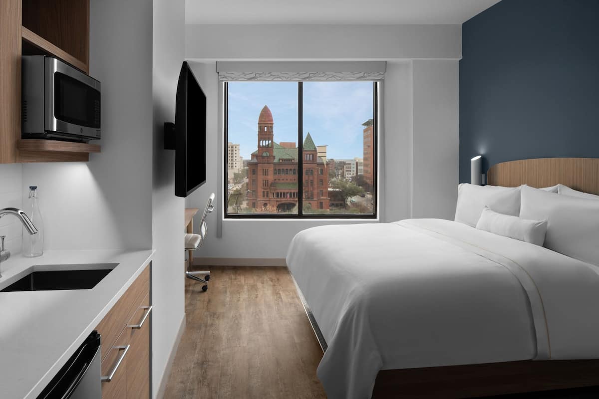 Ein modernes Hotelzimmer mit einem großen Bett, einem flachbildfernseher an der Wand, einer Küchenzeile inklusive Mikrowelle und Blick auf ein historisches Gebäude durch das Fenster.