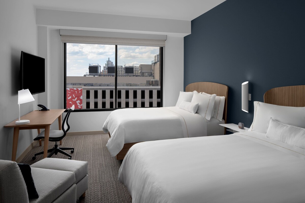 Ein Hotelzimmer mit zwei Betten, einem Schreibtisch mit Stuhl, einem Fernseher und einem großen Fenster mit Blick auf die Stadt.