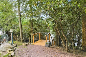 Walk across the wooden bridge to a secluded restroom or shelter surrounded by lush trees and a gravel path.
