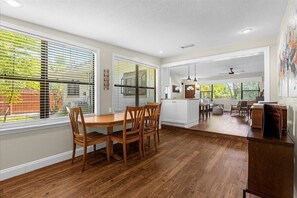 A spacious kitchen with wooden flooring, large windows with blinds, and a dining area with chairs.
