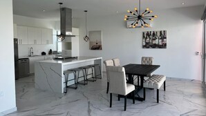 Una cocina moderna con armarios blancos, una isla central con taburetes de bar y un comedor con piso de mármol y una mesa de madera oscura.
