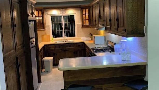A kitchen with dark wooden cabinets, a central island with stools, a window, and a ceiling light. - Apartamento en el centro de la ciudad (La Romana)