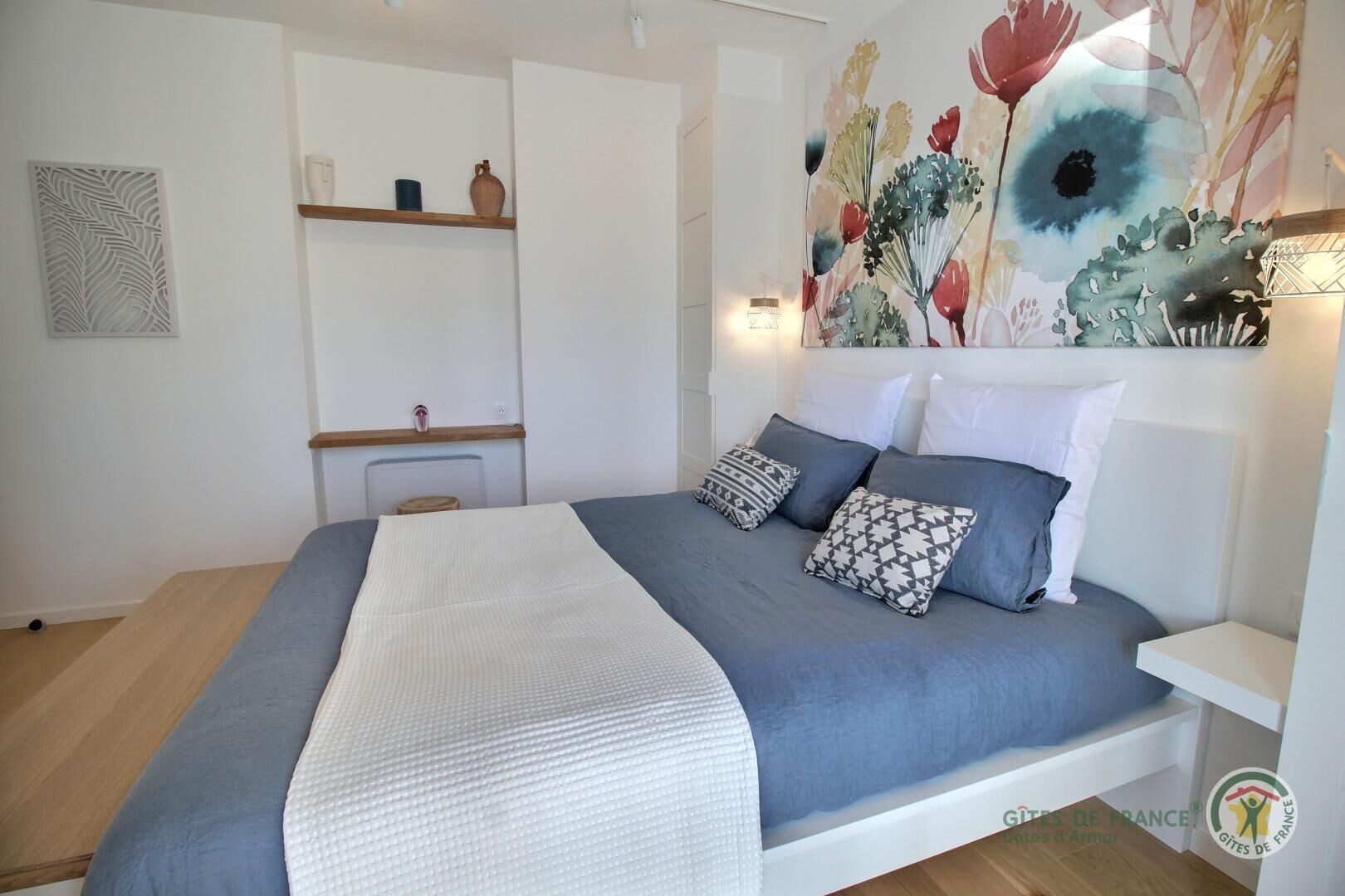 A bedroom with a bed, pillows, a bedside table, a wall-mounted shelf, and a decorative painting.