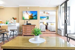 A modern hotel room with a dining area, a TV, a ceiling fan, and a large window. - Waikiki Banyan condo - pool, hot tub & parking (Honolulu)
