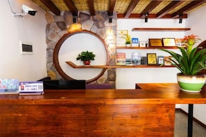 Un comptoir d’accueil avec un plateau en bois, un mur en pierre, un miroir rond et une étagère ornée d’objets encadrés et d’une plante en pot.