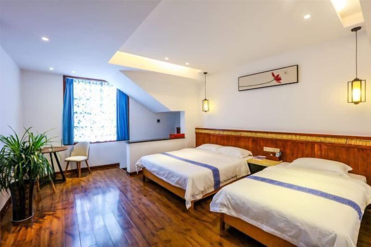 Une chambre d’hôtel avec deux lits, un bureau, une chaise, une plante en pot et une fenêtre avec des rideaux bleus.