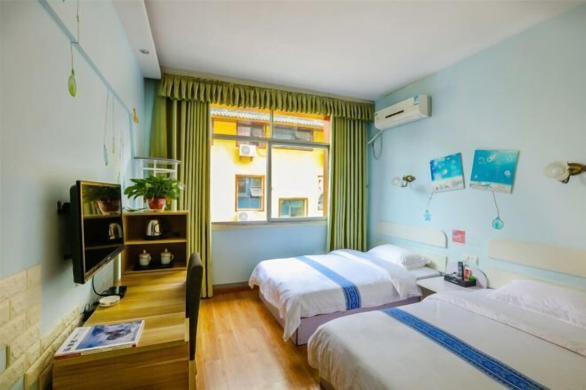 Habitación de hotel con dos camas, un escritorio de madera, una estantería con libros y una planta, una ventana con cortinas y una obra de arte en la pared.