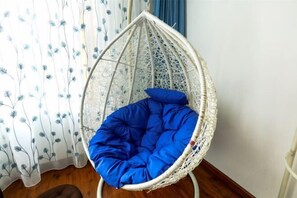Una silla colgante de mimbre blanco con un cojín azul en una habitación con piso de madera y cortina estampada.