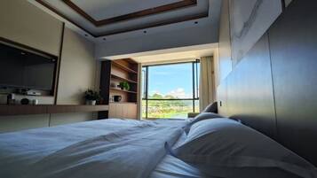 A bedroom with a large bed, a built-in shelf with plants, and a view of a green landscape through an open door.
