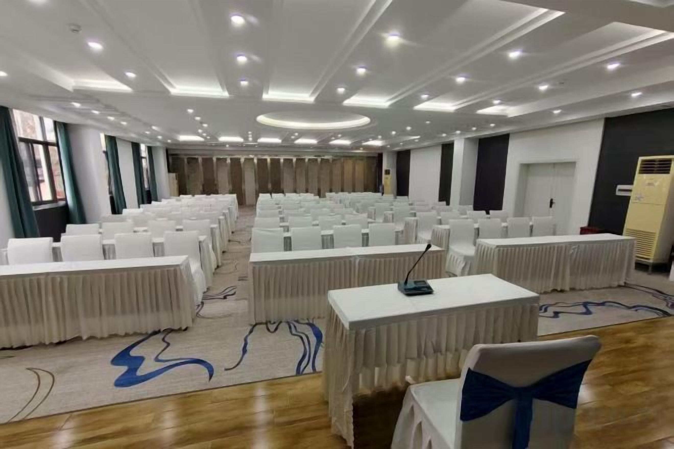A conference room with rows of white tables and chairs, a carpeted floor, and a ceiling with recessed lighting.