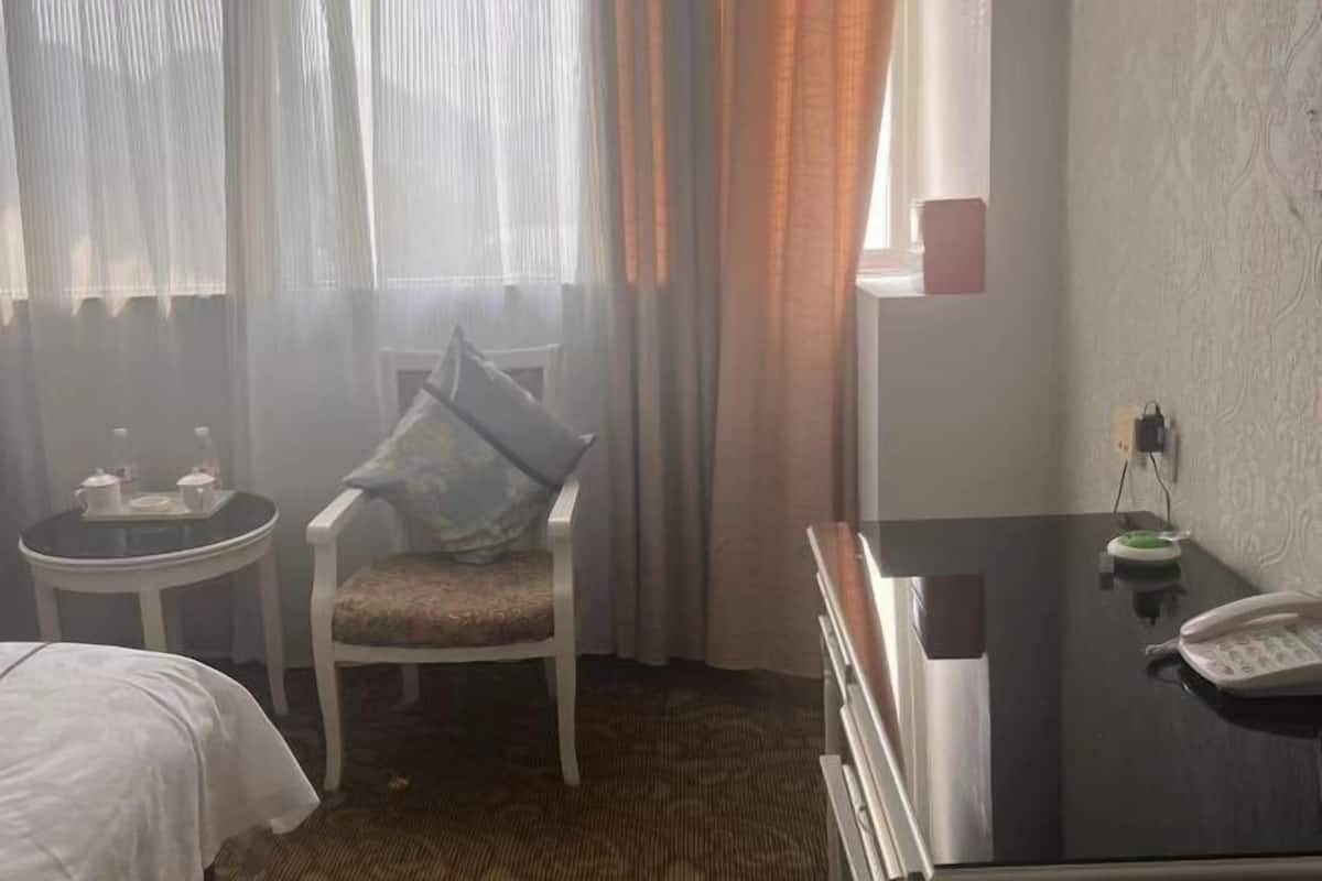 Ein Hotelzimmer mit einem Bett, einem Sessel, einem kleinen Tisch mit Tablett, einem Schreibtisch mit Telefon und einem Fenster mit Vorhängen.